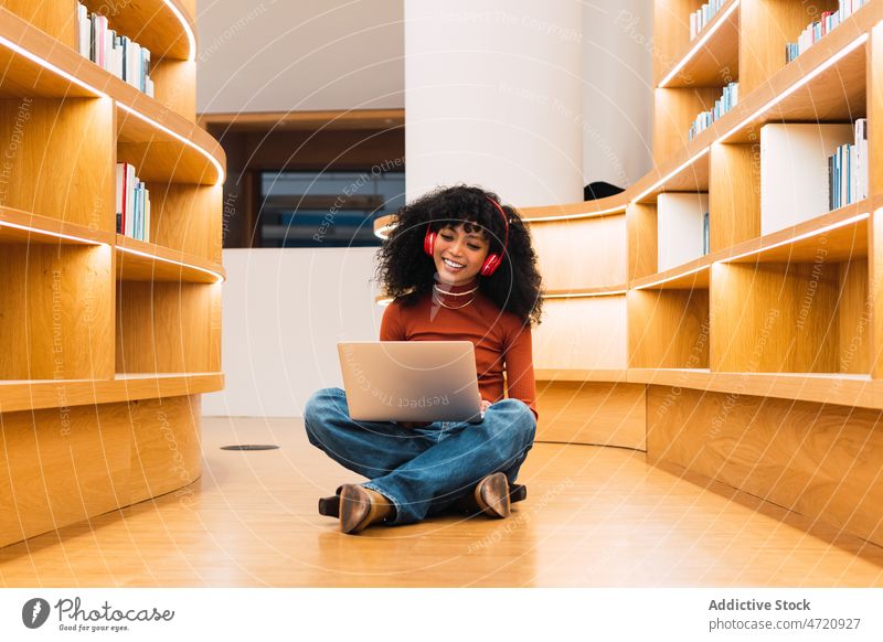 Afroamerikanische Frau benutzt Laptop in Bibliothek zu Musik benutzend meloman Schüler Kopfhörer Lächeln Bücherregal Bücherschrank zuhören Browsen Bildung