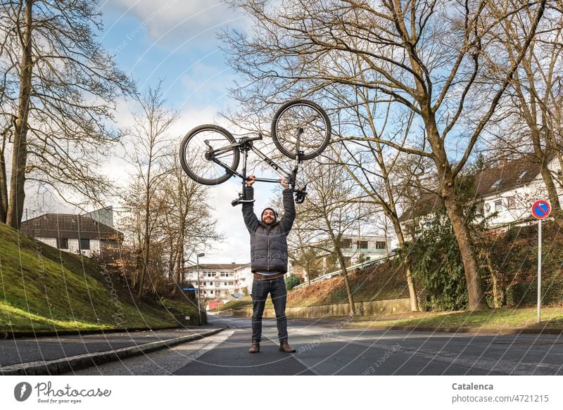 Junger Mann hebt sein Fahrrad hoch Radfahrer urban Grau Tageslicht Bäume Wohngebiet Aktion Fahrradfahren Sport Stadt Winter Straße Person Himmel Wolken Kraft