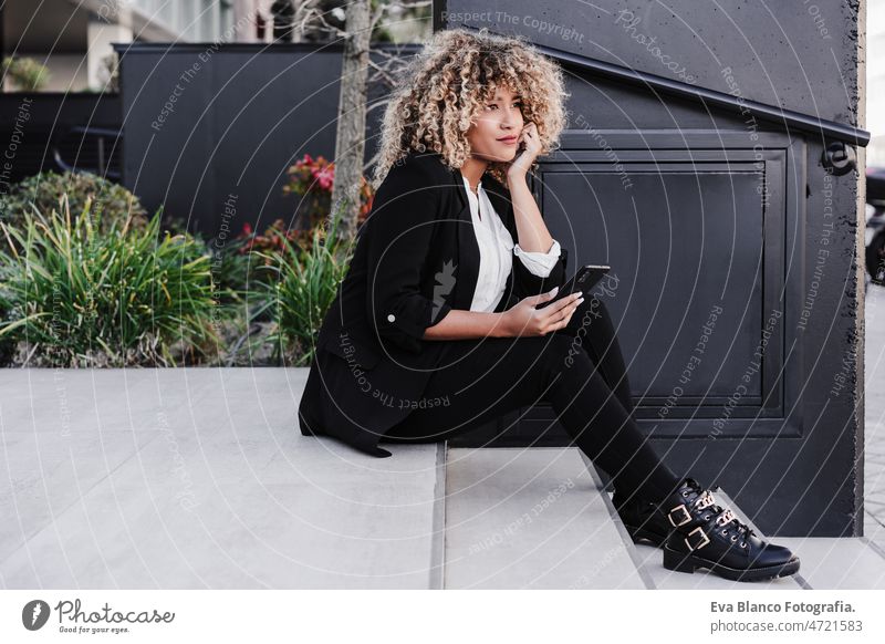 schöne lächelnde Business-Frau mit Handy sitzen auf Treppen in der Stadt. Gebäude Hintergrund Afro-Look hispanisch Geschäftsfrau Großstadt Wolkenkratzer jung