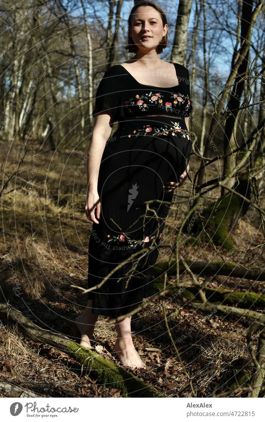 Junge, schwangere Frau steht in schwarzem Umstandskleid barfuß im Wald junge Frau schön Schönheit neues Leben Kleid Schwangerschaftsfoto Bauch Babybauch