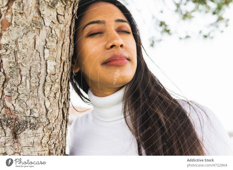 Lächelnde indische Frau in der Nähe eines Baumes Vorschein Straße Großstadt urban Ast feminin Stil Kofferraum Revier lange Haare geschlossene Augen attraktiv