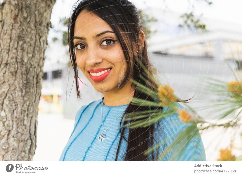 Lächelnde indische Frau in der Nähe eines Baumes Vorschein Straße Großstadt urban Ast feminin Stil Kofferraum Revier lange Haare attraktiv charmant Inhalt