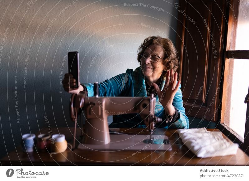Fröhliche Näherin macht Selfie in der Nähe der Nähmaschine Frau Nähen Basteln Smartphone Wellenhand Hobby reif Senior Rentnerin Vorrichtung Faser Instrument