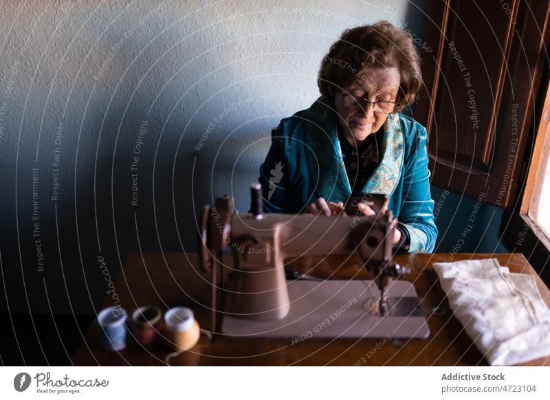 Ältere Frau näht an einer Maschine Nähmaschine Nähen Textil Näherin Basteln Hobby Vorrichtung Faser Instrument Stil Garn Design Raum Handwerkerin Tisch Licht