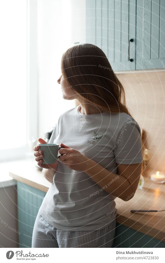 junge Frau trinkt morgens Kaffee aus einer Keramiktasse, lächelt in den neuen Tag, steht in der Küche und schaut zum Fenster Morgen gute Miene zum bösen Spiel