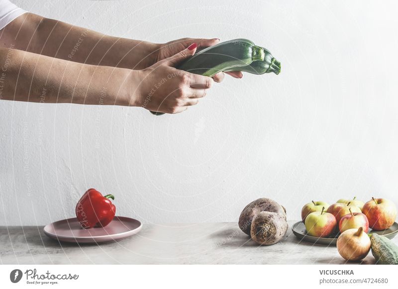 Frau hält zwei grüne Zucchini an einem grauen Küchentisch mit Obst und Gemüse Hände Beteiligung Tisch Früchte weiß Wand Hintergrund Gesunder Lebensstil