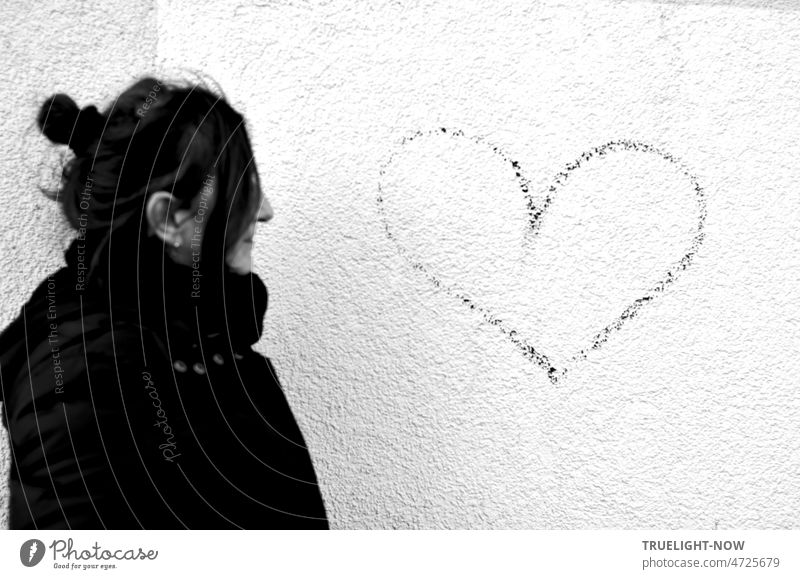 Annäherung: warm eingepackte Frau, das Haar hoch gebunden, bei der Betrachtung von Herz Liebesbotschaft auf weißer Hauswand Profil lange Haare Mantel Schal