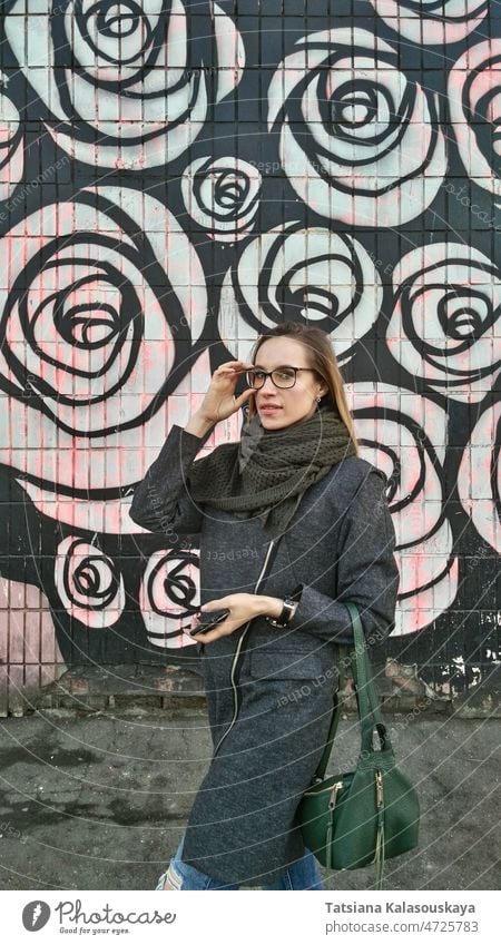Eine junge Frau in einem grauen Mantel und mit grünem Schal rückt eine Brille vor dem Hintergrund einer farbigen Wand zurecht. Model Tasche schön Stil Mädchen