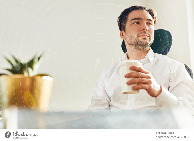 Junger selbstbewusster Geschäftsmann sitzt am Schreibtisch im Büro. Entspannter Mann in weißem Hemd hält eine Tasse Kaffee und schaut weg arbeiten Business