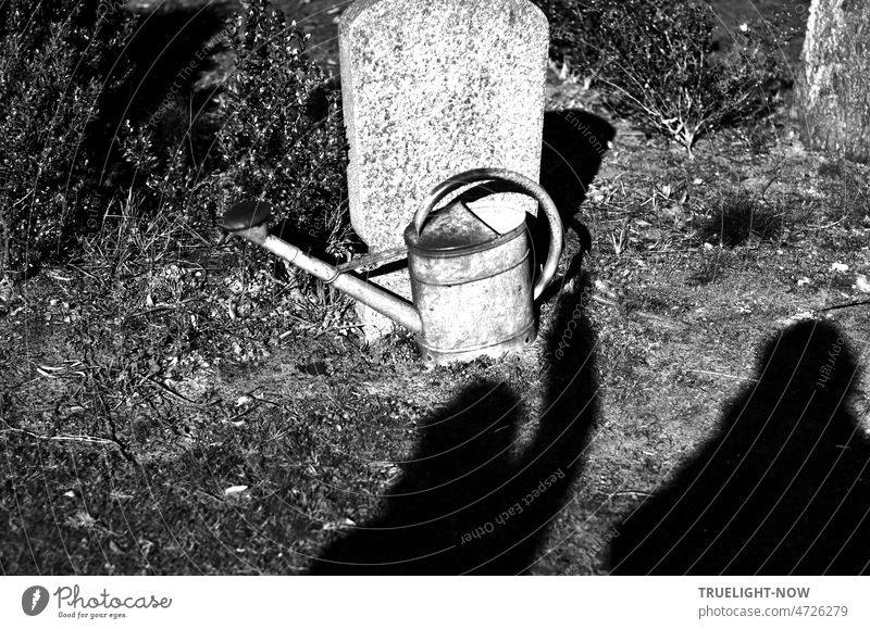 Sonnenschein auf dem Friedhof in schwarz-weiss: zwei Schatten auf der Erde an einem alten Grabstein und ein Schatten-Arm greift nach einer alten Gieskanne, die vor dem Stein steht