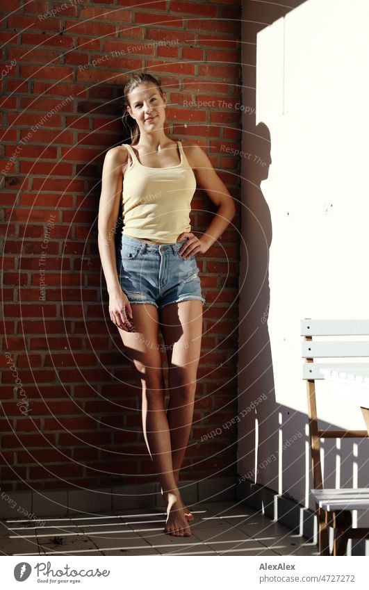 Junge, große, blonde Frau steht gegen eine Backsteinwand gelehnt auf dem Balkon barfuß in der Sonne und lächelt in die Kamera schön schlank ästhetisch sportlich