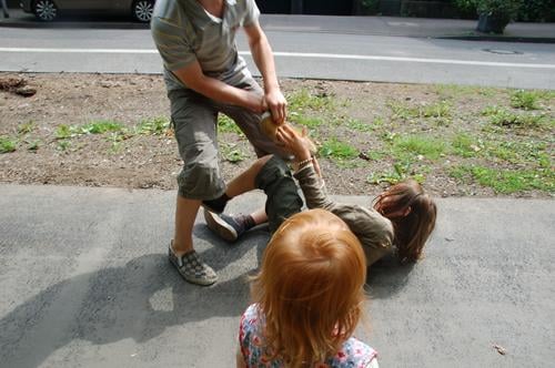 zwei Jungs raufen auf einem Bürgersteig und ein rothaariges Mädchen schaut zu kämpfen Kinder Jungen Kindheit Kinderspiel Soziales Leben Spielen Spaß spielerisch
