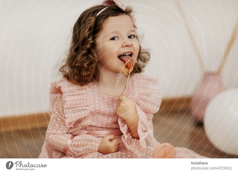 Ein kleines Mädchen in einem rosa Kleid sitzt auf dem Boden und isst einen Lutscher Kind wenig Bonbon Lollipop Essen niedlich süß Kinder und Zucker Baby