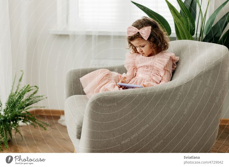Ein kleines Mädchen sitzt auf einem Stuhl und spielt ein Spiel auf ihrem Handy oder schaut Cartoons. Baby Smartphone zuschauend Technik & Technologie Kind