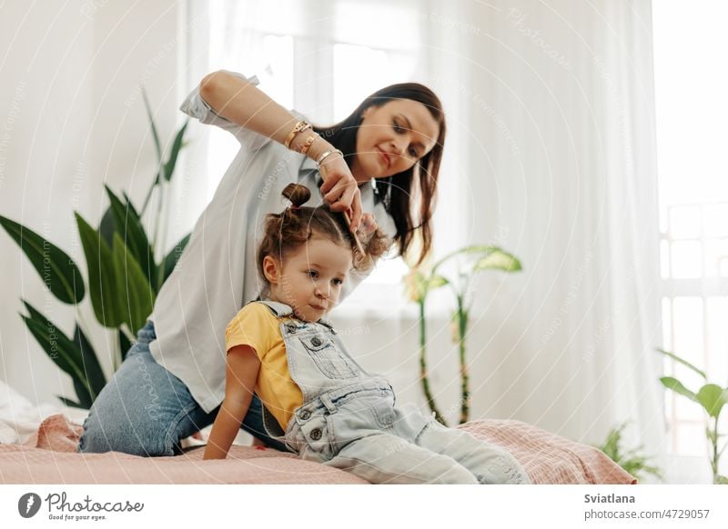 Eine glückliche Mutter kämmt ihrer kleinen Tochter die Haare, während sie morgens im Bett sitzt Baby Kamm Behaarung Familie Glück Sitzen Haarbürste Morgen