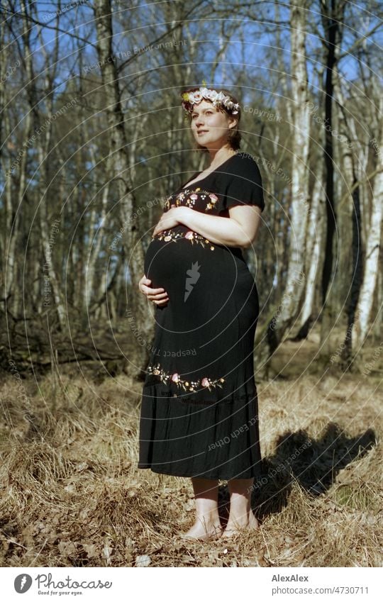 Analoges Ganzkörperportrait einer jungen, schwangeren Frau, die barfuß und mit Blumenkranz im Haar in einem Laubwald steht und sich den Babybauch mit einer Hand hält