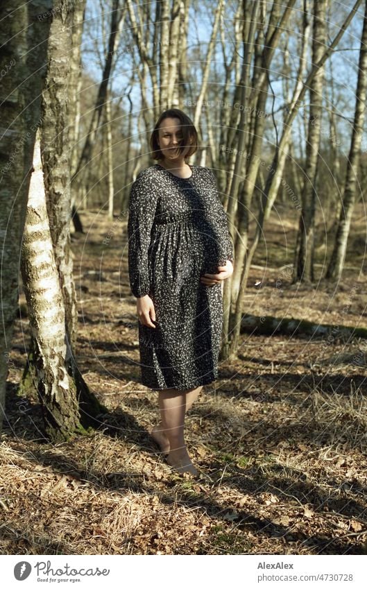 Analoges Ganzkörperportrait einer jungen, schwangeren Frau, die barfuß in einem Laubwald steht und sich den Babybauch mit einer Hand hält Lächeln kraftvoll Gras