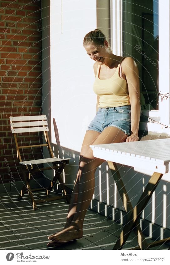 Junge, große, blonde Frau lehnt gegen ein Fenster auf dem Balkon barfuß in der Sonne und lächelt - analoges Portrait schön schlank ästhetisch sportlich