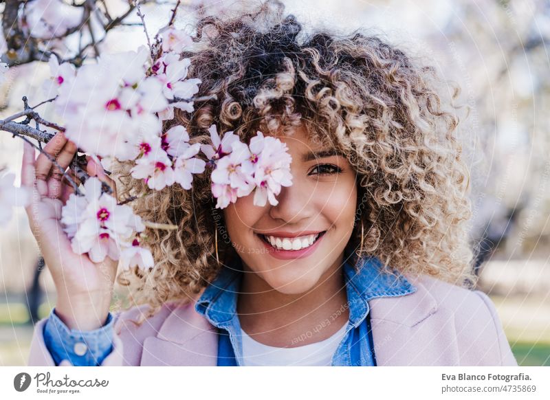 lächelnde hispanische Frau mit geschlossenen Augen im Park genießt sonnigen Tag. Frühling Blumen Hintergrund Tanzen kurvenreich Körperfreundlichkeit Afro-Look