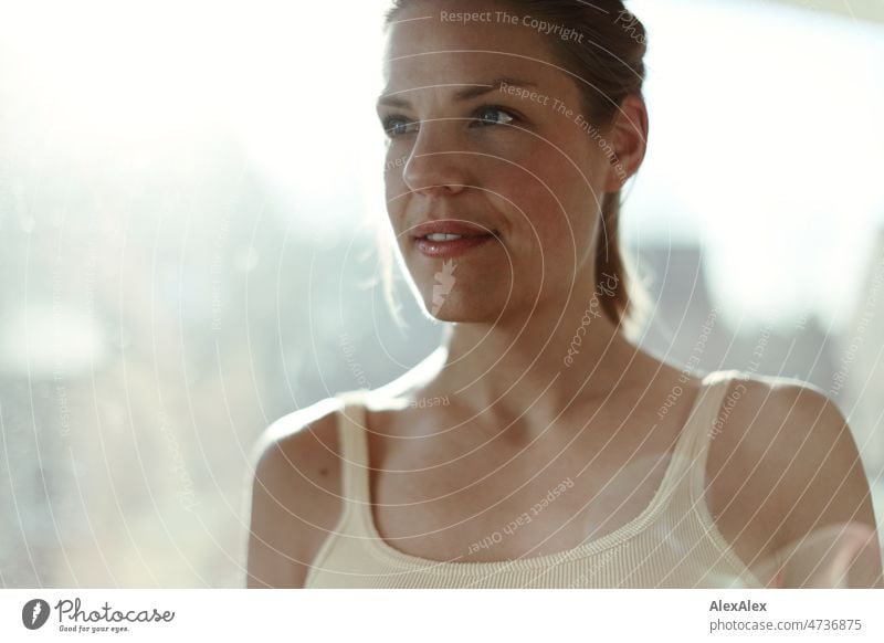 Gegenlicht- Portrait einer jungen, großen, blonden Frau vor einer Fensterscheibe Tanktop freundlich glücklich Zentralperspektive Nahaufnahme Jugendlichkeit