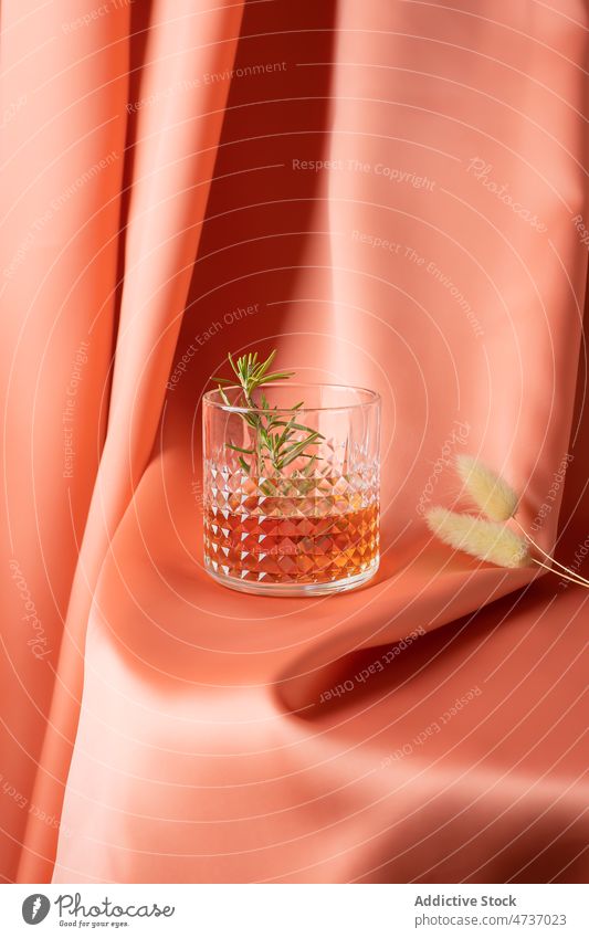 Glas kalter Whiskey mit Rosmarinzweig auf Stoff serviert aromatisch Alkohol elegant Schnaps Gewebe Vorhang Seide Zweig Stillleben Lagurus Getränk Kristalle