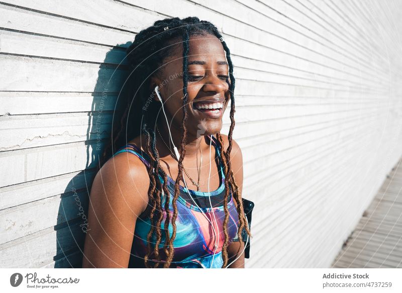 Fröhliche schwarze Frau in Sportkleidung, die Musik über Kopfhörer hört zuhören Fitness Training Wiedergabeliste Freizeit Glück Lachen positiv sportlich