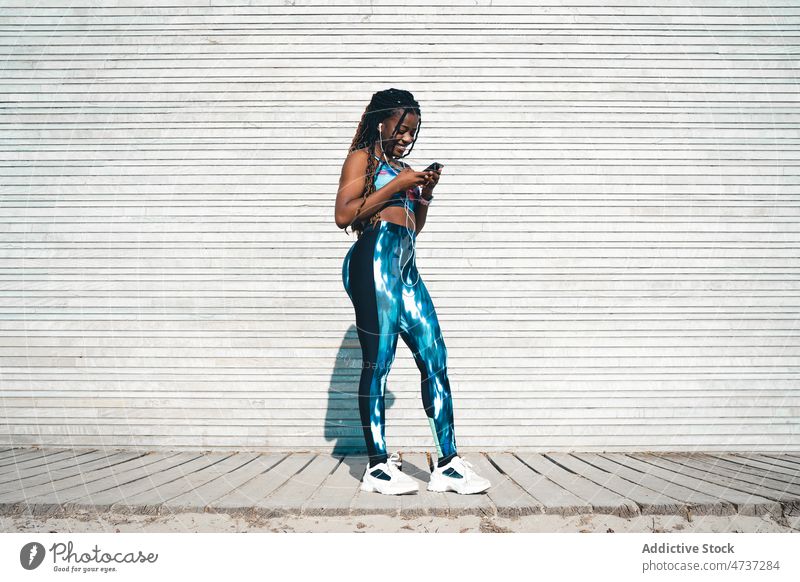 Lächelnde schwarze Frau in Sportkleidung, die ihr Smartphone in der Nähe einer Holzwand benutzt benutzend zuhören Kopfhörer Training Musik Pause Fitness