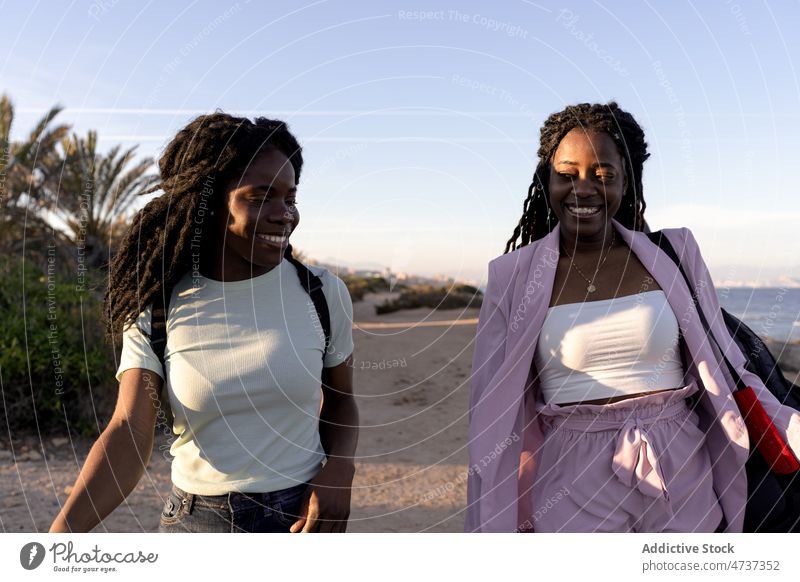 Fröhliche schwarze Frauen, die bei Sonnenuntergang am Strand spazieren gehen Spaziergang Sommer Zusammensein Freund Lifestyle Meeresufer Abend ethnisch