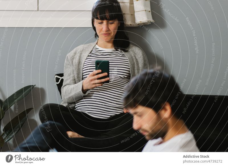 Schwangere Frau surft in der Wohnung ihres Mannes auf dem Smartphone Paar digital Süchtige benutzend Anschluss Internet lesen Ehefrau Wohnzimmer schwanger Gerät