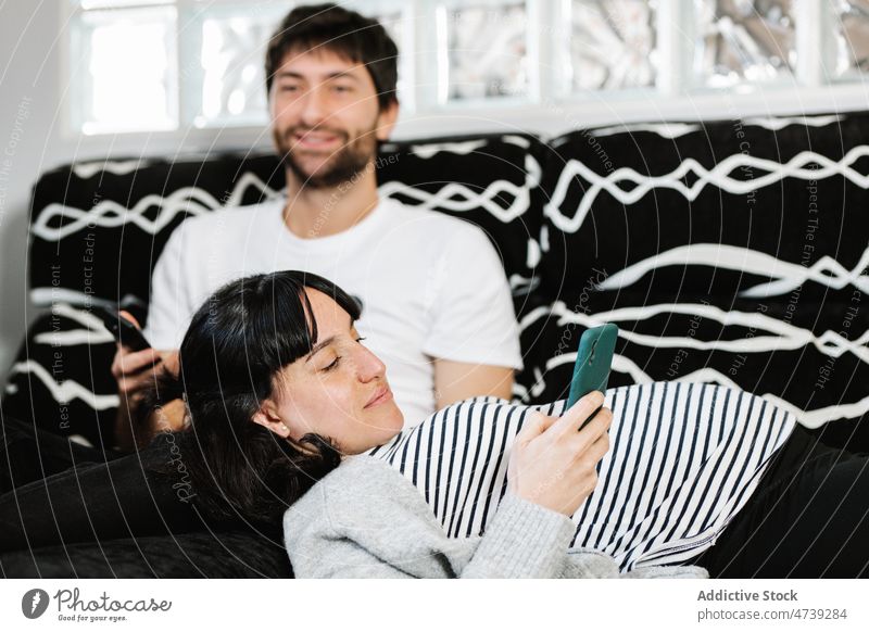 Junger hispanischer Mann, der auf dem Sofa neben seiner schwangeren Frau fernsieht und ein Smartphone benutzt Paar ruhen FERNSEHER Zeit verbringen Lügen