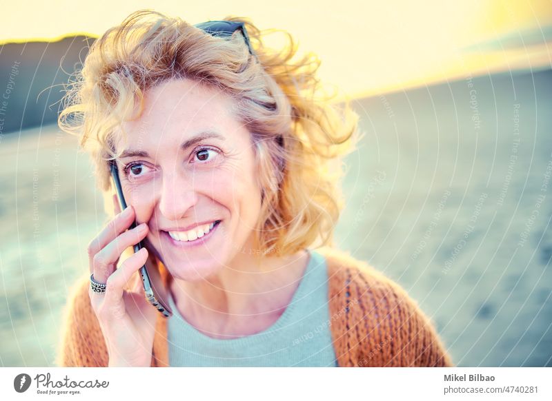 Junge reife blonde kaukasische Frau im Gespräch mit einem Mobiltelefon im Freien an einem Strand. Lifestyle-Konzept. Frauen Mobile Mitteilung Handy