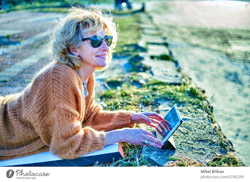 Junge reife blonde kaukasische Frau, die sich an einem sonnigen Tag mit einem Tablet im Freien entspannt. Lifestyle Porträt Frauen Gesundheit verbunden