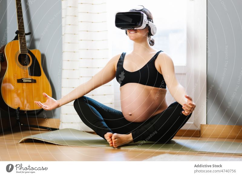 Lächelnde schwangere Frau meditiert mit VR-Brille zu Hause Yoga meditieren Cyberspace Virtuelle Realität Schwangerschaft heimwärts Schutzbrille Dehnung heiter