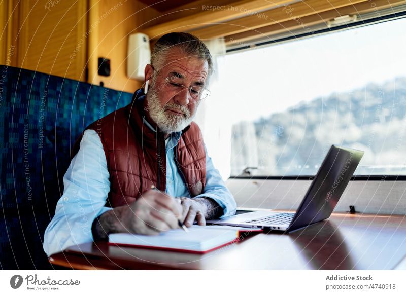 Älterer Mann, der in einem Wohnwagen in der Nähe eines Laptops in einen Notizblock schreibt Wohnmobil rv online Autoreise reif Senior Notebook schreiben