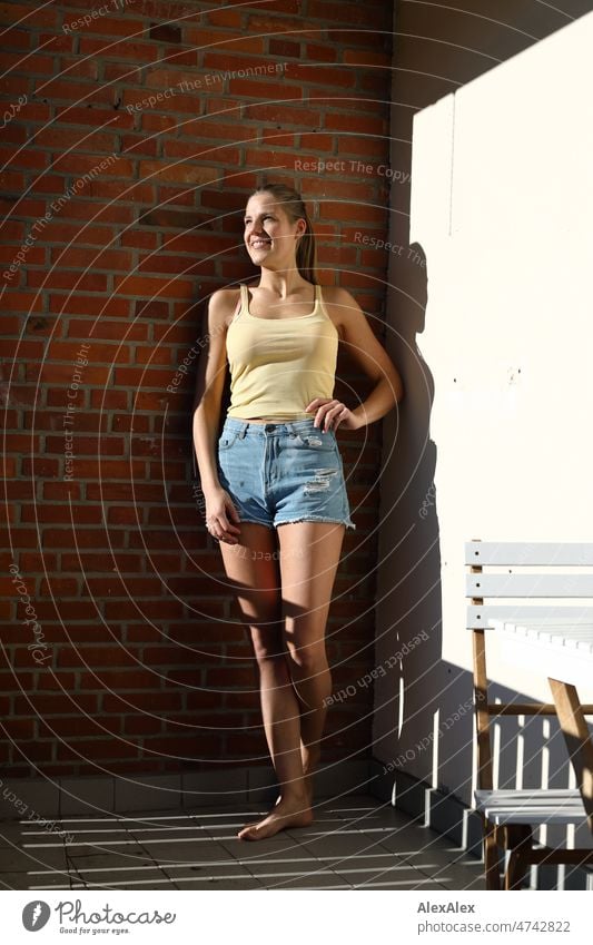 Junge, große, blonde Frau steht gegen eine Backsteinwand gelehnt auf dem Balkon barfuß in der Sonne Selbstbewußt Persönlichkeit Lifestyle Jugendlichkeit