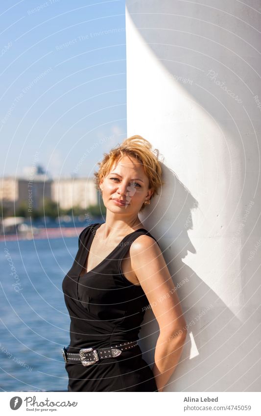 Porträt eines jungen Mädchens mit Kurzhaarschnitt. Ein Mädchen an einem sonnigen Tag posiert auf dem Damm in einem schwarzen Kleid. Porträt eines Mädchens blond