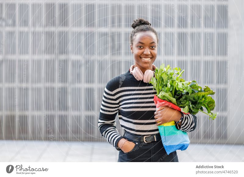 Erfreute schwarze Frau mit Grün an der Wand Kopfhörer Straße Suppengrün Rote Beete Lächeln gesunde Ernährung organisch Vitamin Metallwand Gesundheit Tasche