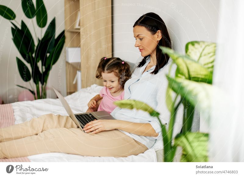 Eine junge Mutter arbeitet am Laptop oder sieht sich mit ihrer kleinen Tochter im Schlafzimmer Videos an. Gemeinsame Zeit, online arbeiten Bett Familie