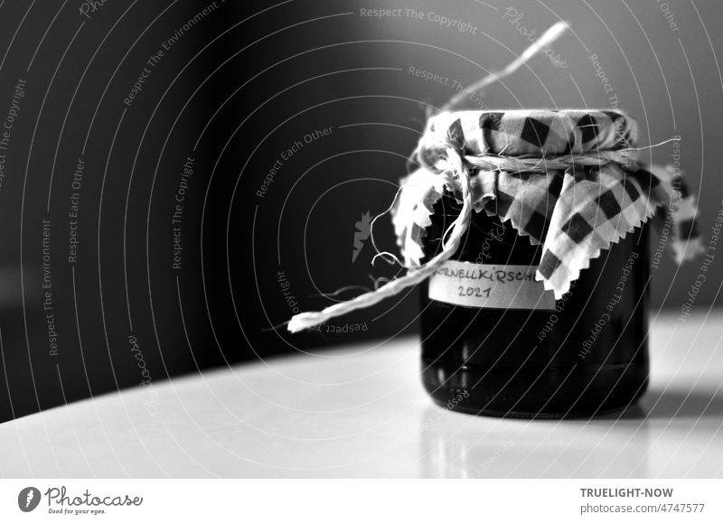 Marmeladenglas mit handgeschriebenem Etikett Kornell Kirsche Konfiture 2021, Dekoration Karo Muster Stoff mit gezacktem Rand, gebunden mit Bindfaden auf weisser runder Tischplatte vor dunklem Hintergrund