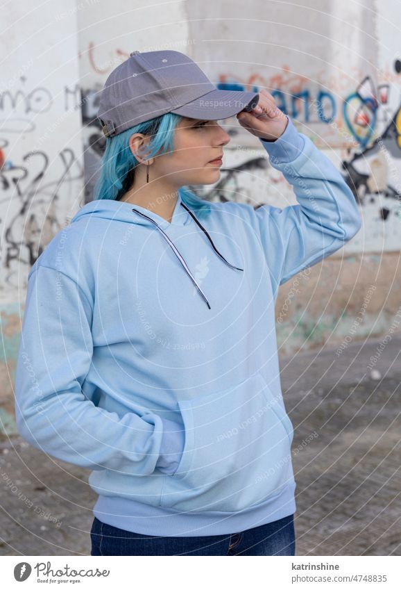 Blauhaariges Teenager-Mädchen in blauem Kapuzenpulli und Baseballmütze, das an einer Graffiti-Wand steht hell-blau Attrappe Verschlussdeckel blauhaarig