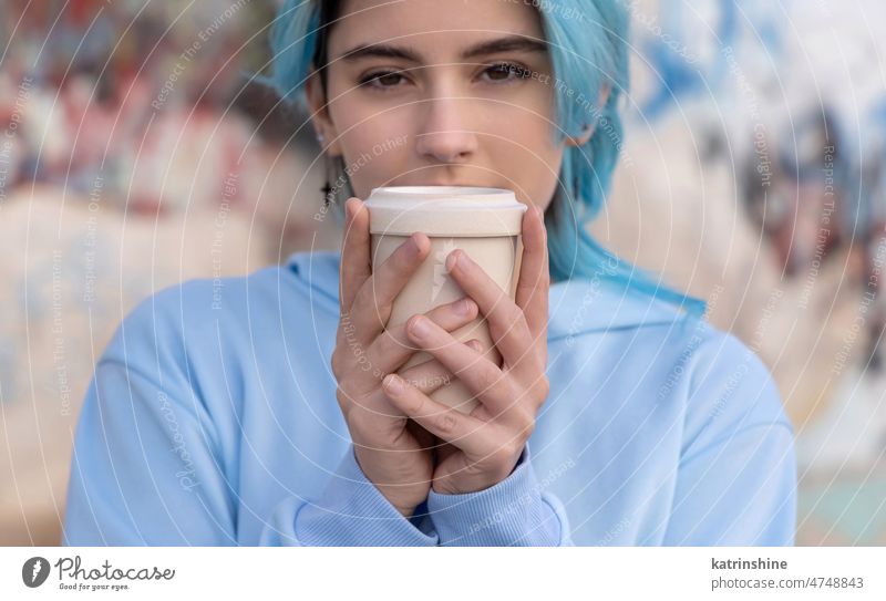 Blauhaariges Teenager-Mädchen in hellblauem Oversize-Kapuzenpulli in der Nähe einer Graffiti-Wand mit Kaffee zum Mitnehmen hell-blau Attrappe Übergröße Tasse