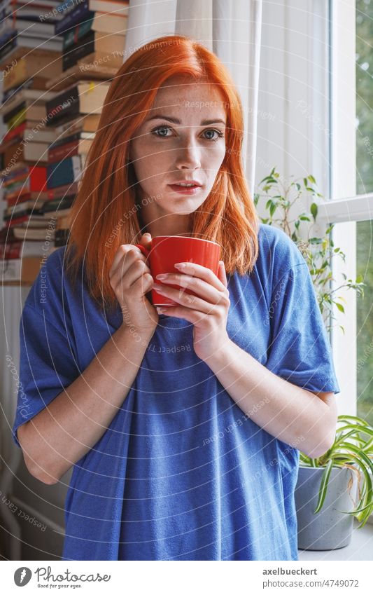 junge Frau steht mit Kaffeetasse am Fenster in ihrer Wohnung Junge Frau zuhause echte Menschen Tasse Kaffeetrinken Heißgetränk Getränk genießen stehen gemütlich