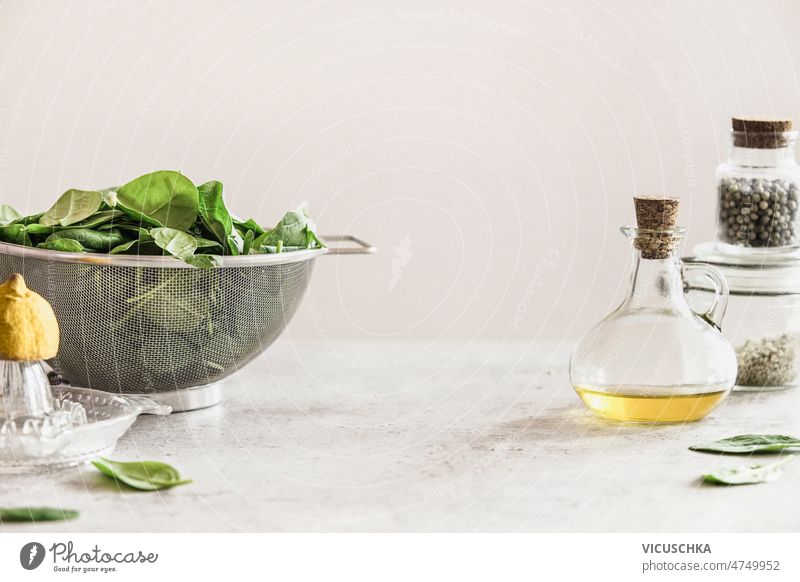 Lebensmittel Hintergrund mit frischem Spinat Blätter in Sieb, Olivenöl in Glasflasche und Zitruspresse auf weißen Küchentisch an der Wand Hintergrund. Gesundes Kochen zu Hause. Vorderansicht.