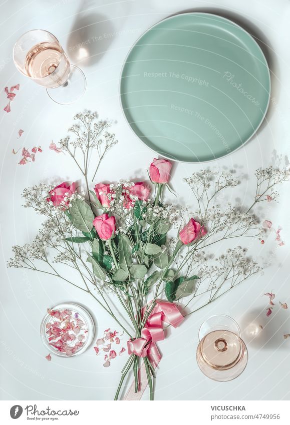 Blumenstrauß mit rosa Rosen, leerer Teller und Rosenwein in Gläsern im Sonnenlicht Ästhetik flache Verlegung Weingläser weißer Hintergrund Draufsicht