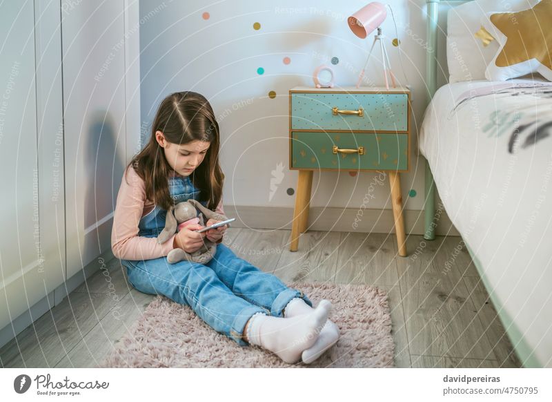 Kleines Mädchen, das ein Handy benutzt, während sie ihr Stofftier auf dem Boden sitzend umarmt wenig benutzend spielen Umarmung gefüllt Spielzeug Sitzen Stock