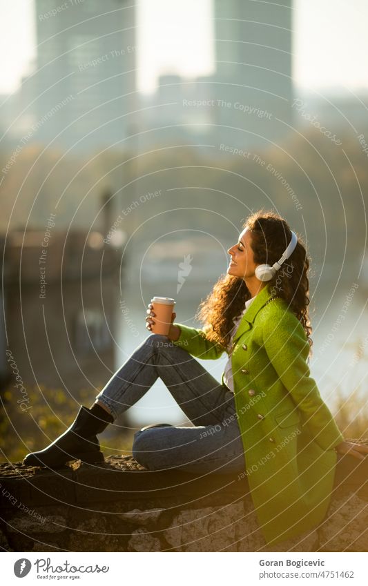 Hübsche junge Frau hört Musik mit Smartphone und Kaffee zum Mitnehmen am Fluss an einem sonnigen Herbsttag hübsch attraktiv Kaukasier im Freien schön