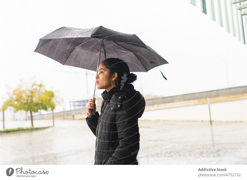 Ethnische Frau mit Regenschirm steht bei Regen auf der Straße bedeckt wow Wunder Reaktionen u. Effekte urban behüten Saison Überraschung Jacke Wetter Großstadt