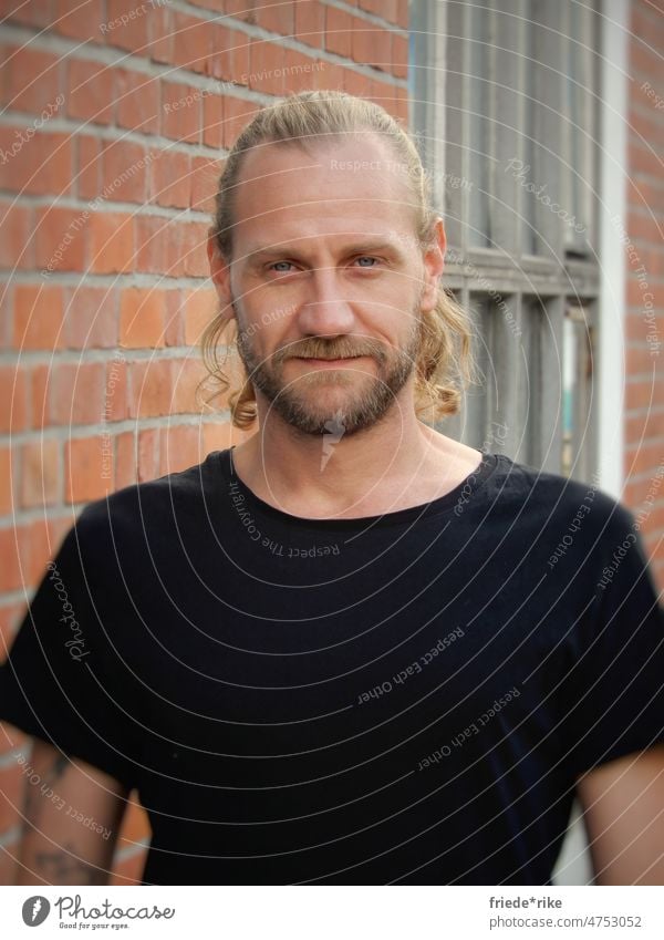 Mann vor Backsteinwand Porträt Bart Locken Fabrik schwarz T-Shirt Tattoo lange Haare blond blaue augen lächelnd geheimnisvoll