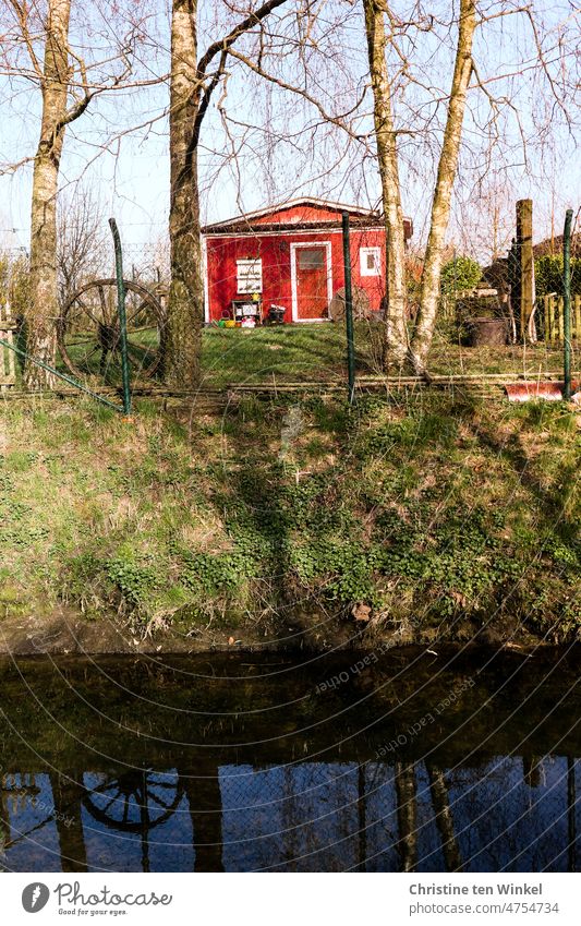 Eine rote Hütte in einem kleinen Garten direkt am Wasser Gartenhaus Wochenendhaus Idylle Spiegelung Birken Bäume Rad grün Gärtchen Schrebergarten