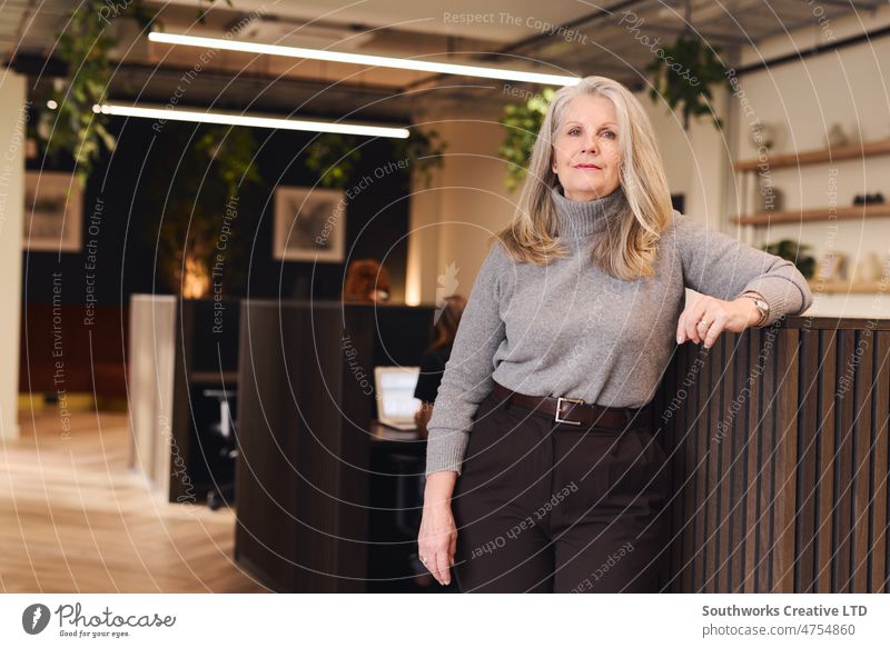 Porträt einer älteren weißen Geschäftsfrau mit konzentriertem Ausdruck, die sich an eine Bürotrennwand in einem modernen Coworking Space lehnt Geschäftsinhaber